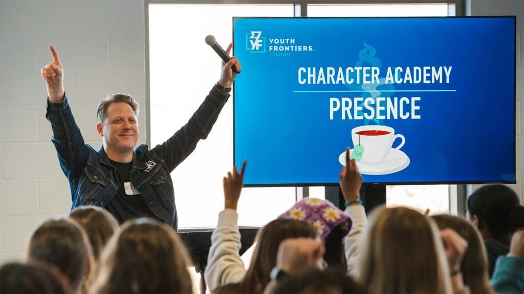 A smiling Youth Frontiers facilitator is standing in front of a crowd of students with arms raised and fingers pointing upwards. Next to him is a tv screen with a keynote slide that reads "Character Academy Presence" with a picture of a cup of tea against a blue background.