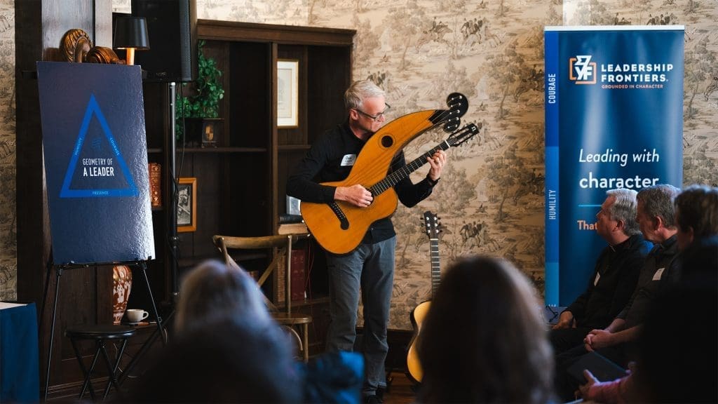 A man standing between two geometry of a leader signs plays a harp guitar to a captivated audience
